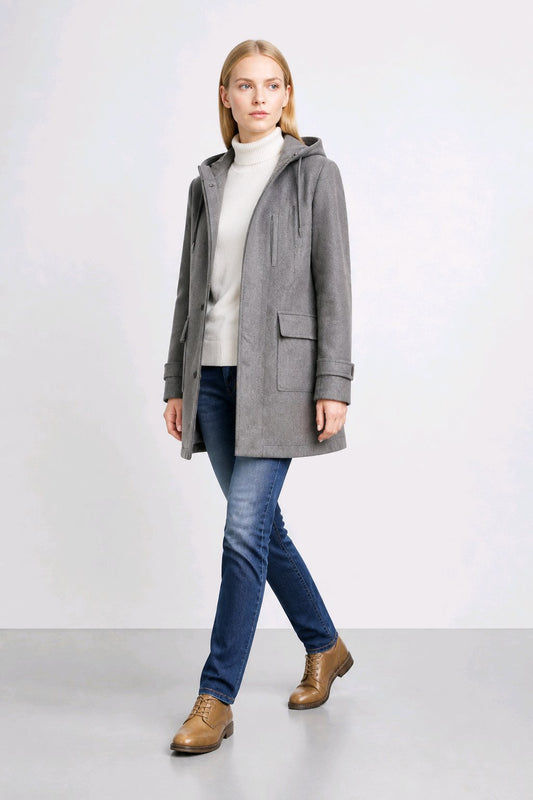 A woman modeling a gray hooded coat over a cream turtleneck sweater, blue jeans, and brown leather shoes, standing against a light gray studio background.