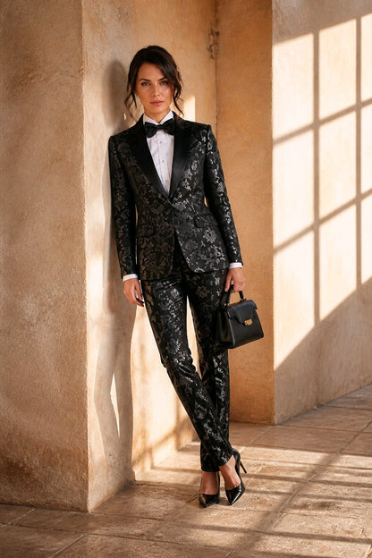 A woman models a black and gold patterned tuxedo suit with a white shirt and black bow tie, standing in a sunlit stone corridor and holding a small black handbag.