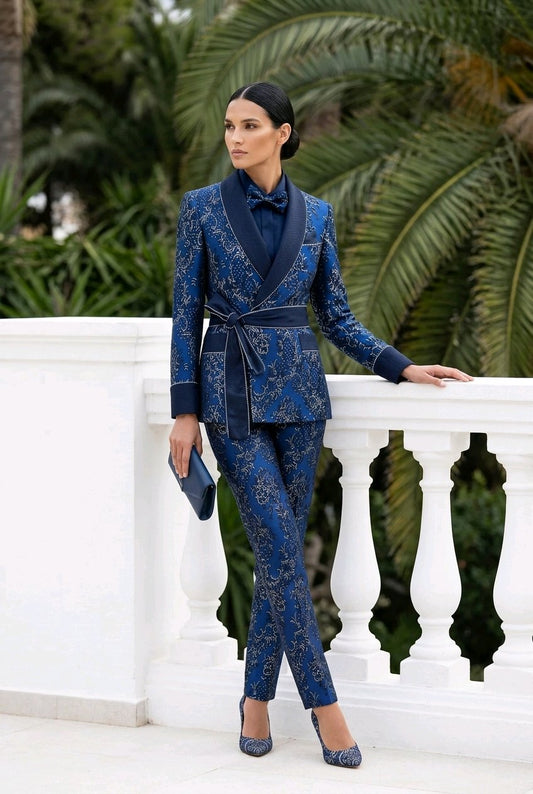 A woman models a blue and gold patterned two-piece suit with a shawl collar blazer and matching trousers, standing on a white terrace with palm trees in the background.