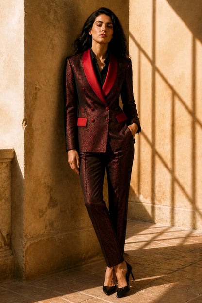A woman models a dark burgundy and black textured suit with a vibrant red shawl collar and matching red pocket flaps, standing in a sunlit stone corridor.