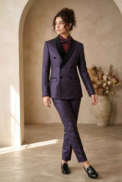 A woman models a dark purple double-breasted suit with a subtle glittery texture, black peak lapels, and a matching purple bow tie, set against a warm beige architectural background with arches.