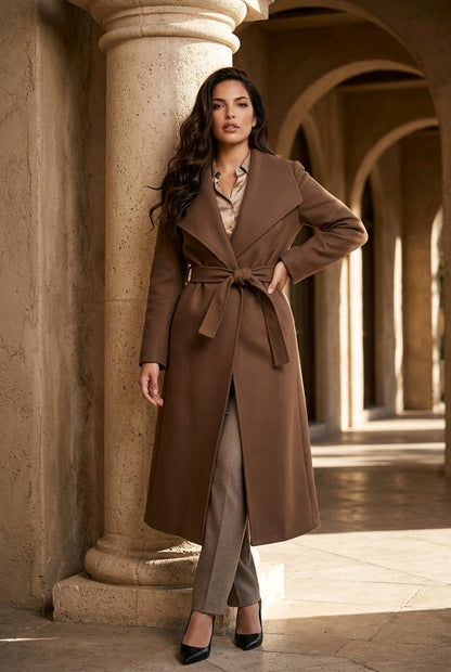 A woman models a long brown belted coat in an elegant stone colonnade setting. She is wearing taupe trousers and black heels.