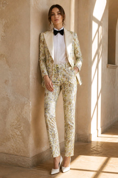 A woman models a matching floral print blazer and trouser set with cream satin lapels, styled with a white shirt, black bow tie, and white pointed-toe heels in a sunlit architectural setting.