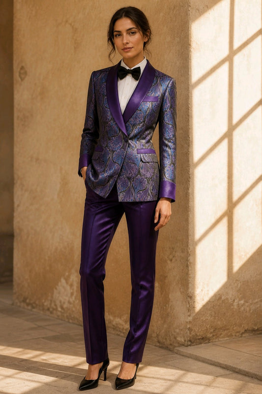 A woman models a purple patterned tuxedo jacket with satin purple shawl lapels, paired with matching purple trousers, a white shirt, and a black bow tie, standing in a sunlit stone corridor.