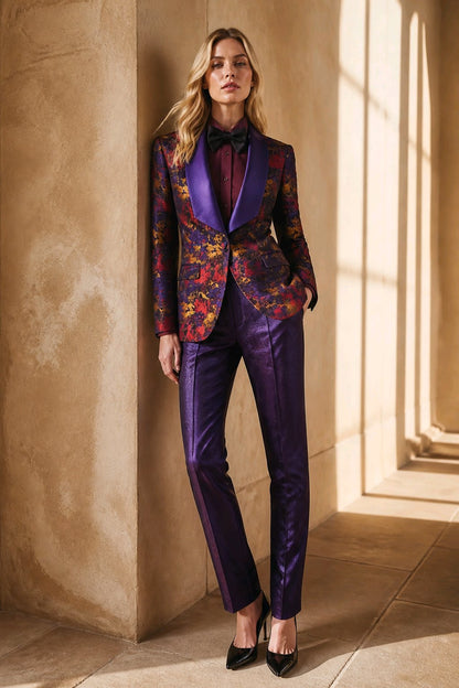 A woman models a purple satin tuxedo suit with a multicolor floral jacquard pattern on the jacket, paired with a black bow tie and black heels, standing in a sunlit stone corridor.
