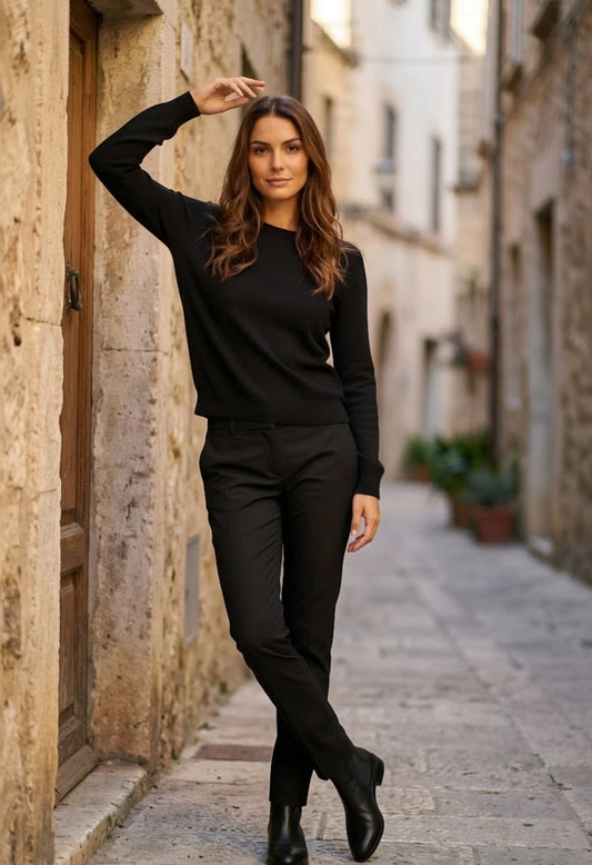 A woman posing in a black long-sleeve crewneck top and black trousers, standing in a narrow stone alleyway.