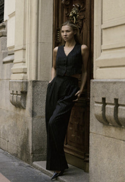 A woman standing in front of a building entrance wearing a black and white pinstripe waistcoat and trousers with no visible brand markings