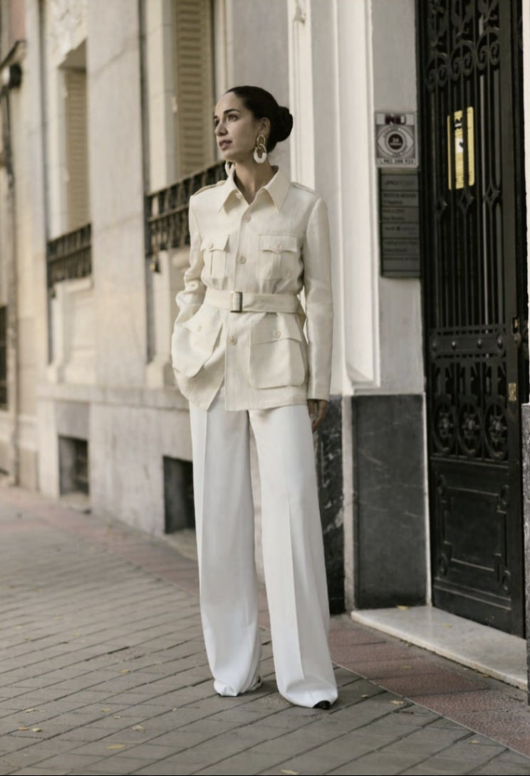 A woman standing on a sidewalk wearing an ivory herringbone safari jacket with five buttons and white trousers.