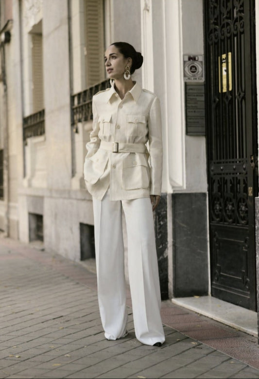 A woman standing on a sidewalk wearing an ivory herringbone safari jacket with five buttons and white trousers.