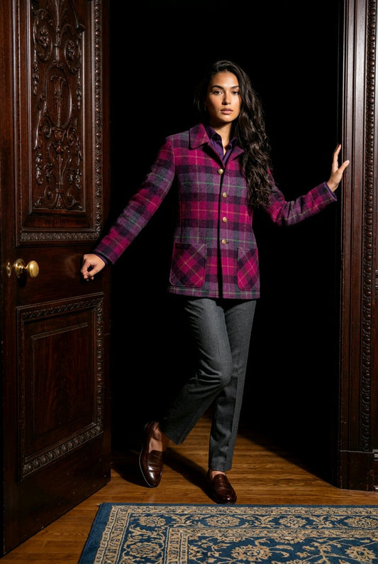 A woman stands in a doorway wearing a magenta and dark gray plaid button-up shirt jacket, dark gray trousers, and brown loafers, with a blue patterned rug on the floor.