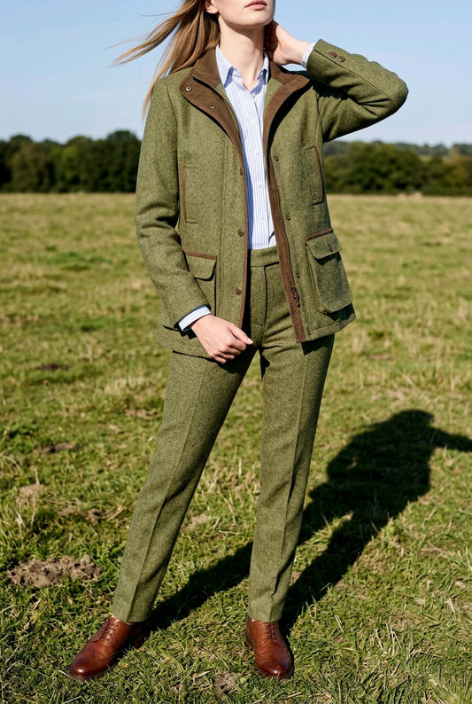 A woman stands in a grassy field wearing an olive green tweed suit with brown corduroy-style lapels and pockets, a light blue shirt, and brown leather shoes.