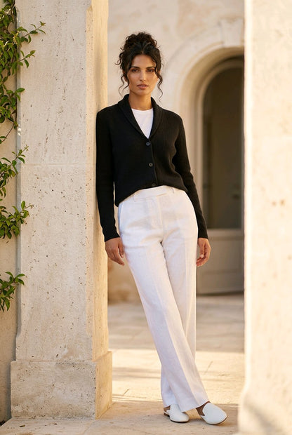 A woman stands in a stone archway wearing a black cardigan over a white top, white wide-leg trousers, and white shoes.