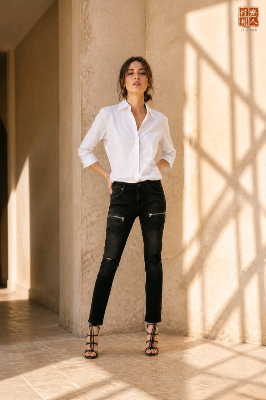 A woman stands in a sunlit architectural corridor wearing a white button-up shirt, black skinny jeans with zip details, and strappy black sandals. A red square logo appears in the top-right corner.