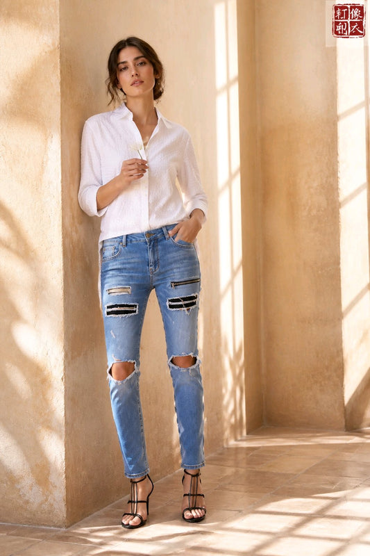 A woman stands in a sunlit beige interior wearing a white button-up shirt, light blue distressed skinny jeans with ripped knees, and black strappy high heels.