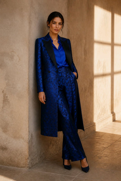 A woman stands in a sunlit interior wearing a luxurious dark blue patterned tailoring set with a long coat, matching trousers, and a bright blue blouse, paired with black heels.