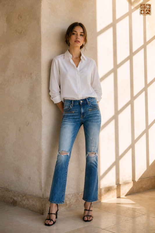 A woman stands in a sunlit interior wearing a white button-up shirt, blue distressed jeans, and black strappy heels, leaning against a textured beige wall.