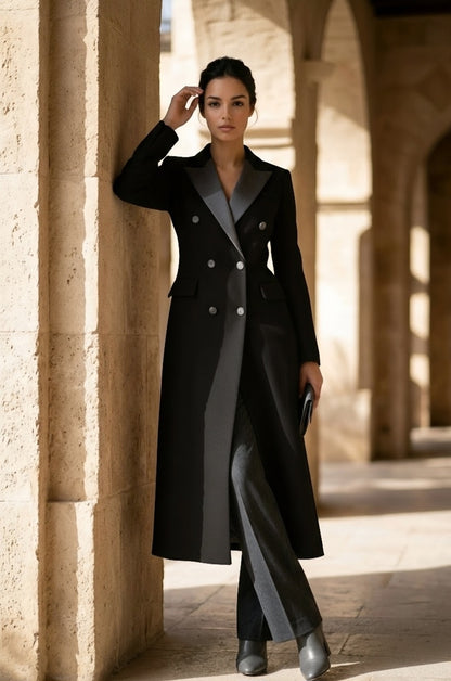 A woman stands in a sunlit stone corridor wearing a long black double-breasted coat over a gray dress and gray trousers, holding a black clutch.
