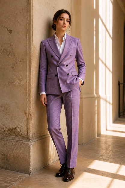 A woman stands in a sunlit stone corridor wearing a tailored lavender-purple double-breasted suit with a white shirt and dark brown leather shoes.
