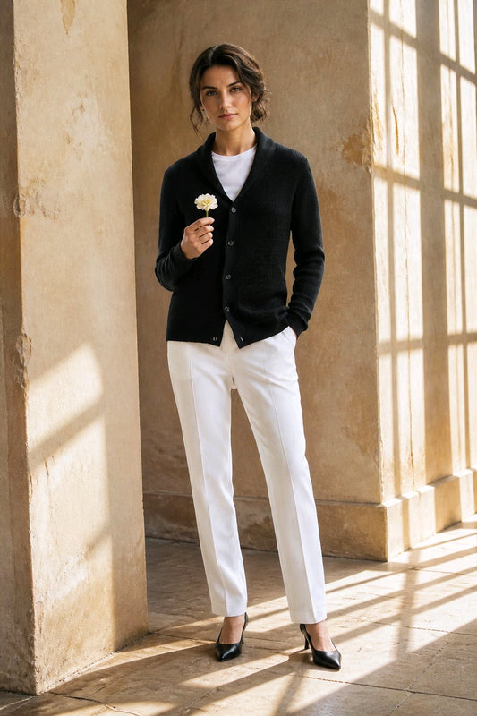 A woman stands in a sunlit stone interior wearing a black button-front cardigan over a white top, white trousers, and black pointed heels, while holding a small white flower.
