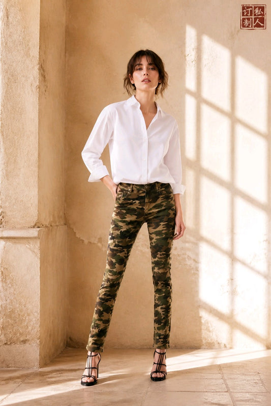 A woman stands in a sunlit stone interior wearing a white button-up shirt, slim-fit camouflage pants, and black strappy heels. A red square logo appears in the top-right corner.