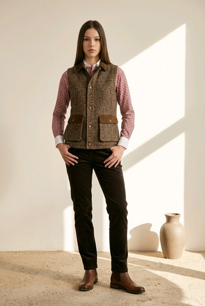 A woman stands in a sunlit studio wearing a brown tweed vest over a red-and-white gingham shirt, dark trousers, and brown boots. A beige ceramic vase sits on the floor against a cream wall.