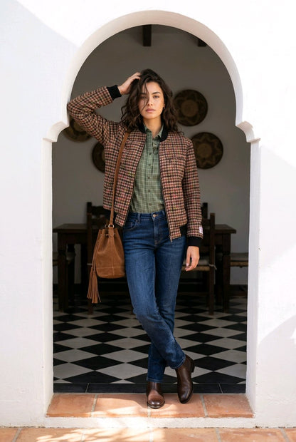 A woman stands in a white arched doorway wearing a brown and beige plaid blazer over a green shirt, blue jeans, and brown boots, with a brown leather tassel shoulder bag.