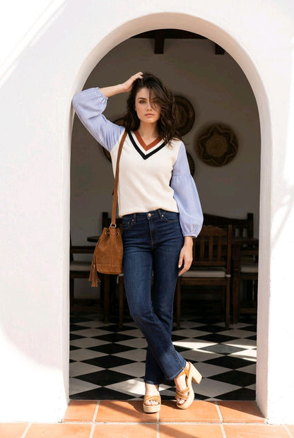 A woman stands in a white arched doorway wearing a cream knit sweater vest with a black and navy V-neck trim over light blue balloon sleeves, paired with dark blue jeans, a brown leather shoulder bag, and beige platform sandals.