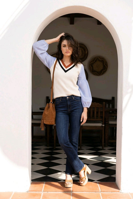 A woman stands in a white arched doorway wearing a cream knit sweater vest with a black and navy V-neck trim over light blue balloon sleeves, paired with dark blue jeans, a brown leather shoulder bag, and beige platform sandals.