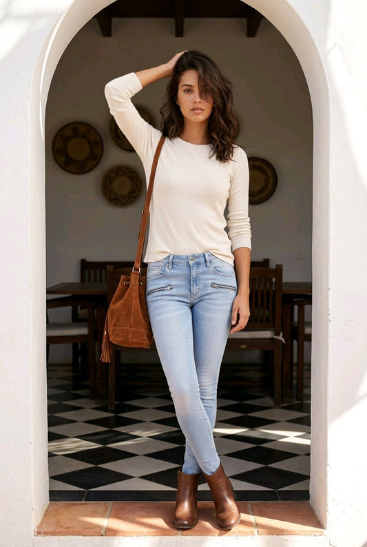 A woman stands in a white arched doorway wearing a cream long-sleeve top, light blue skinny jeans, and brown ankle boots, with a brown shoulder bag. The background shows a warm interior with wooden furniture and a black-and-white checkered floor.