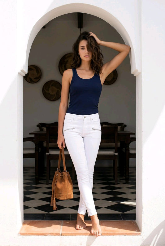 A woman stands in a white arched doorway wearing a navy blue tank top and white skinny jeans, holding a tan leather bucket bag.