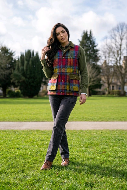 A woman stands on a grassy park path wearing a colorful plaid quilted vest over an olive green jacket, dark gray jeans, and brown shoes.