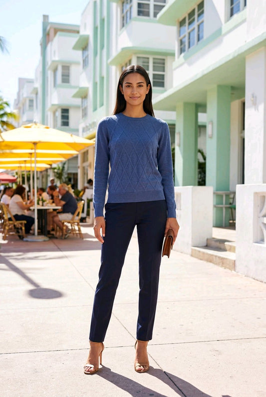 A woman stands on a sunny sidewalk wearing a blue textured knit sweater and dark navy pants, with a beachfront building and outdoor cafe seating in the background.