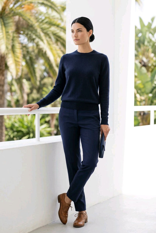 A woman stands on a white balcony wearing a dark navy long-sleeve knit sweater and matching navy trousers, with brown leather shoes and holding a black clutch. Palm trees are visible in the background.