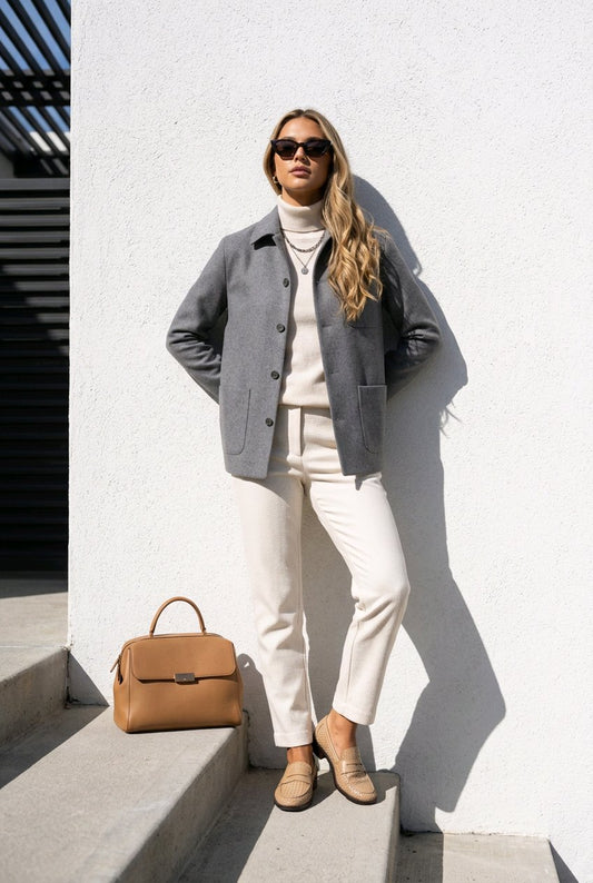 A woman stands on concrete steps against a white wall wearing a gray button-front jacket over a cream turtleneck, cream pants, and tan loafers. A tan handbag rests beside her on the steps.
