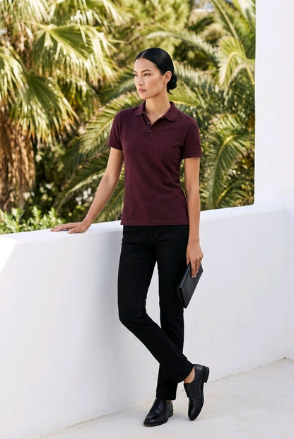 A woman stands outdoors against a white wall with palm trees behind her, wearing a short-sleeve polo shirt in deep burgundy and black pants. She holds a black clutch and wears black shoes.