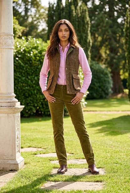 A woman stands outdoors in a garden wearing a pink button-up shirt, a brown tweed vest, olive green corduroy trousers, and brown shoes.