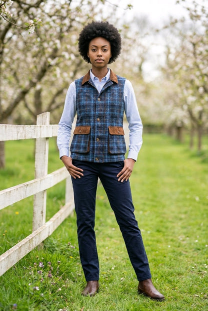 A woman stands outdoors in a grassy field with flowering trees and a white fence, wearing a blue plaid hunting vest with brown leather-like trim over a light blue shirt and dark navy trousers.