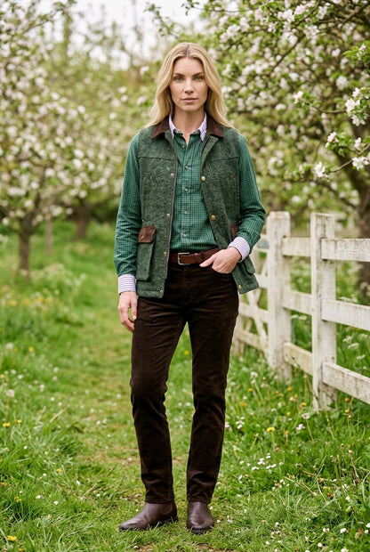 A woman stands outdoors in a grassy orchard with white blossoms, wearing a green textured jacket over a green shirt, dark brown corduroy trousers, and brown boots.