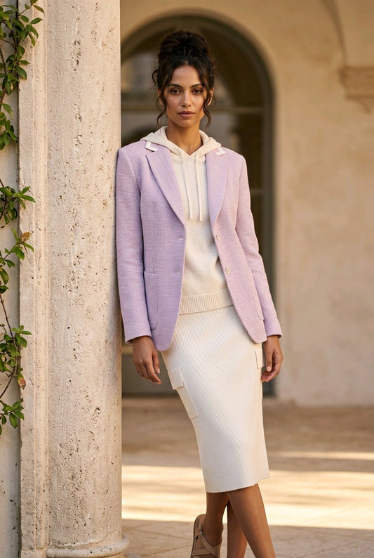 A woman stands outdoors in front of a stone archway wearing a light lavender textured blazer over a cream hoodie and a matching cream midi skirt.