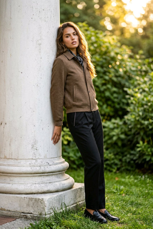 A woman stands outdoors leaning against a large white stone column, wearing a brown suede jacket, black trousers, and black loafers. The background features green foliage and warm golden sunlight.