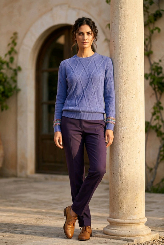 A woman stands outdoors next to a stone column wearing a periwinkle blue cable-knit sweater, dark purple trousers, and brown leather shoes.