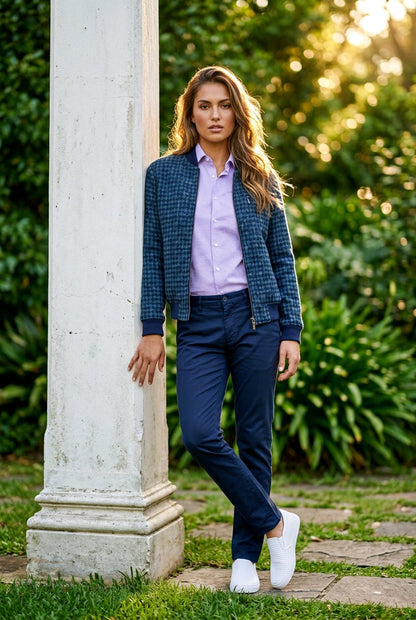 A woman stands outdoors next to a white stone column, wearing a navy blue plaid blazer over a light purple button-up shirt, navy trousers, and white sneakers.