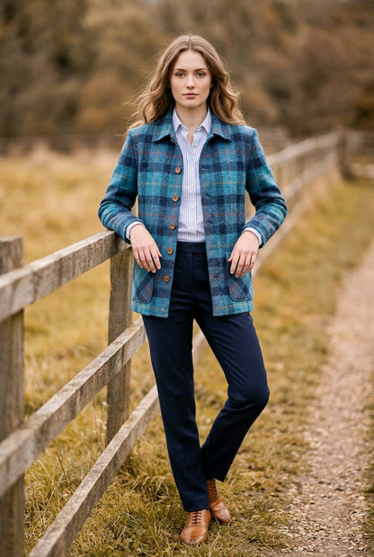 A woman stands outdoors on a dirt path beside a wooden fence, wearing a blue and teal plaid button-up jacket over a light blue shirt, navy trousers, and brown leather shoes.
