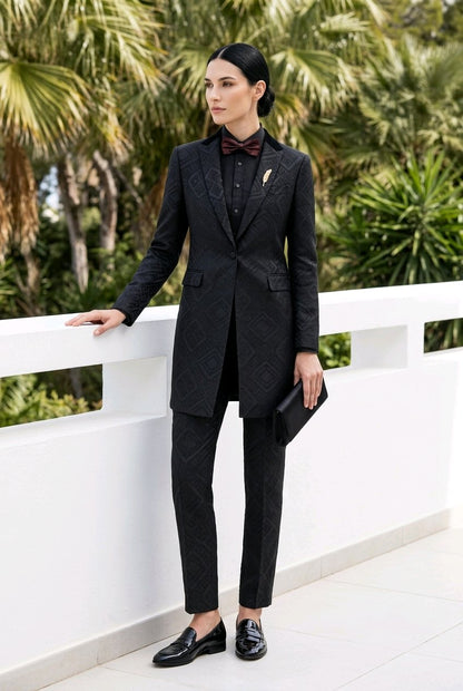 A woman stands outdoors on a white terrace wearing a tailored black patterned suit with a blazer, slim trousers, black loafers, a black shirt, and a dark bow tie. Palm trees are visible in the background.
