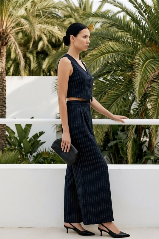 A woman stands outdoors wearing a dark navy pinstriped two-piece set consisting of a sleeveless V-neck crop top and matching wide-leg trousers, paired with black heels and holding a black clutch bag.