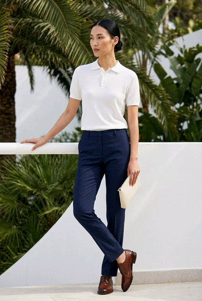 A woman stands outdoors wearing a white short-sleeve polo shirt and navy blue tailored trousers, with brown dress shoes and a light beige clutch, posed in front of a white wall and palm trees.