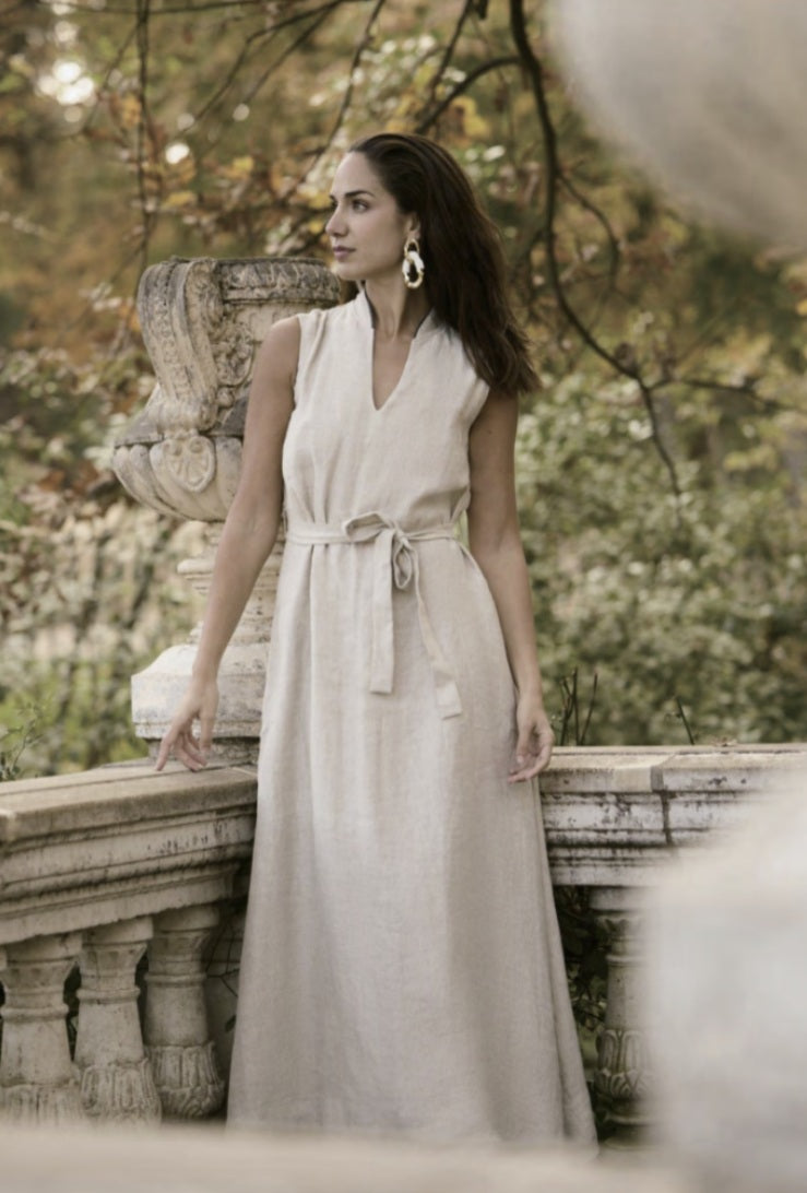 A woman wearing a beige linen full dress with a V-neck, sleeveless top, and belt waist, standing outdoors.