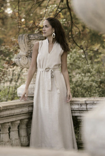 A woman wearing a beige linen full dress with a V-neck, sleeveless top, and belt waist, standing outdoors.