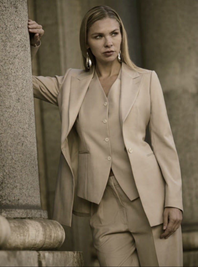 A woman wearing a beige single-breasted, three-piece trouser suit with a wide peaked lapel and straight flapless pockets.