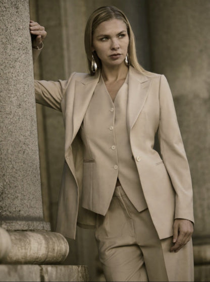 A woman wearing a beige single-breasted, three-piece trouser suit with a wide peaked lapel and straight flapless pockets.